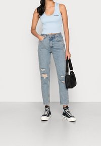 Ljusblå kort topp med texten "BNO JEANS", tillsammans med ljus slitna denimjeans och svarta högstövlar, hållande en svart väska.