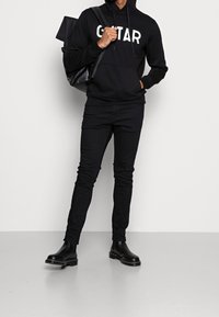 Sudadera negra con la palabra "GUITAR" en letras blancas, combinada con jeans negros ajustados y botas negras de tobillo, sosteniendo una mochila negra.