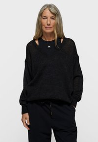 Zwarte, loszittende trui met een V-hals en lange mouwen, gemaakt van een zachte stof met een licht doorschijnende textuur. Gedragen over een zwart topje.