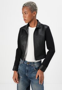 Giacca corta in pelle nera e tessuto con zip frontale, dotata di colletto rialzato e maniche aderenti. Abbinata a jeans in denim blu.
