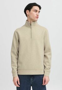 Beige kvart-zip genser laget av teksturert stoff med lange ermer og en avslappet passform. Kombinert med mørkeblå jeans for kontrast.