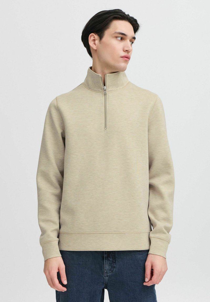 Beige kvart-zip genser laget av teksturert stoff med lange ermer og en avslappet passform. Kombinert med mørkeblå jeans for kontrast.