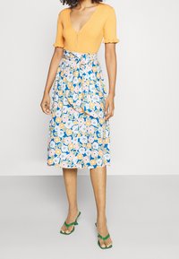 Haut orange à côtes avec des manches courtes et un décolleté en V profond, associé à une jupe fluide bleue ornée d'un motif floral en rose et jaune. Sandales vertes.