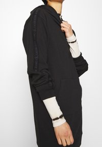 Schwarzes Hoodie-Kleid mit großer Kapuze, Fronttasche und Calvin Klein-Branding entlang des Ärmels. Der Stoff wirkt glatt und hat eine lockere Passform.
