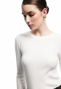 Heller, langärmeliger Pullover mit Kabelstrickmuster und rundem Halsausschnitt. Glatte Textur mit dezenten Rippdetails. Trägt dazu eine dunkle Hose.