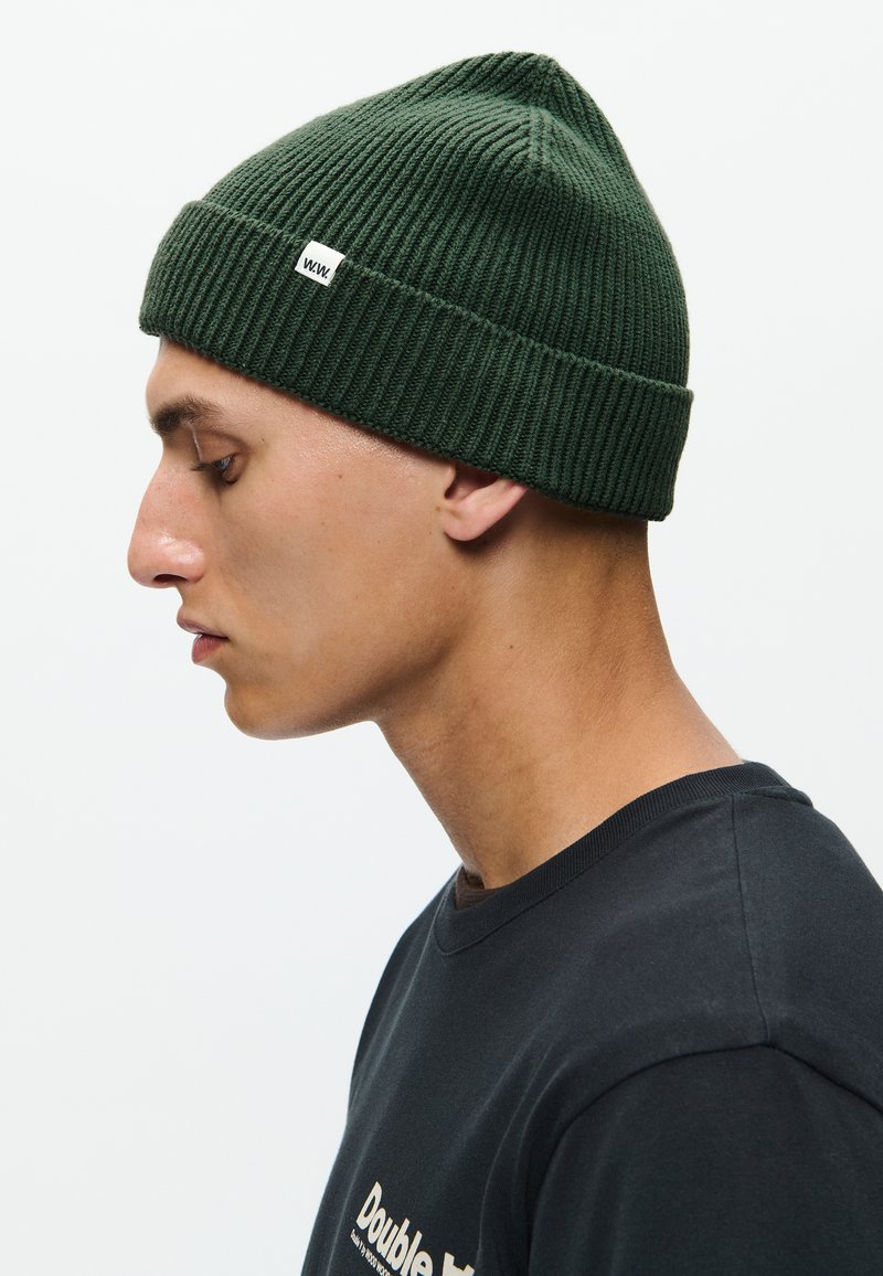 Groene ribgebreide beanie met een piek aan de bovenkant en een klein wit logolabel. Dicht tegen het hoofd gedragen, wat de strakke pasvorm en textuur benadrukt.