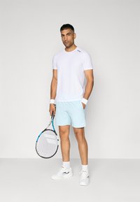 T-shirt atlético branco e calções azul-claro. O modelo segura uma raquete de ténis, vestindo sapatilhas brancas e faixas para os pulsos. Design simples e moderno.