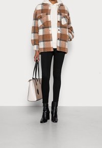 Chemise à carreaux en flanelle marron, blanche et grise, superposée sur un sweat à capuche crème ; associée à un jean skinny noir et des bottes à talons noires. Porte un sac à main beige.