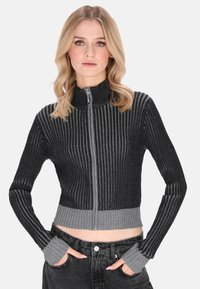 Fekete és szürke bordázott zipzáras pulóver, magas gallérral és hosszú ujjakkal, amelyek hüvelykujj-aljzatokkal rendelkeznek, rövidített fazonnal és kontrasztos mintázattal.