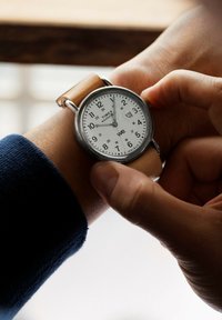 Montre Timex avec un boîtier rond en acier inoxydable, un cadran blanc, des chiffres noirs et un bracelet en cuir marron. Le réglage de la couronne est visible.