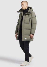 Jachetă căptușită de un verde măsliniu deschis, cu glugă, detalii cu fermoar și o patch cu logo. Asortată cu pantaloni negri și adidași de culoare deschisă.