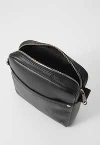 Bolso bandolera de cuero negro con logo en relieve, cierre superior de cremallera y textura suave. Presenta un bolsillo frontal y acentos de hardware en plata.