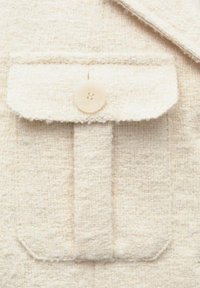 Cremfarbener Jackenstoff mit strukturierter Webart. Verfügt über eine aufgesetzte Tasche und einen runden, vierlochigen Knopf oben.