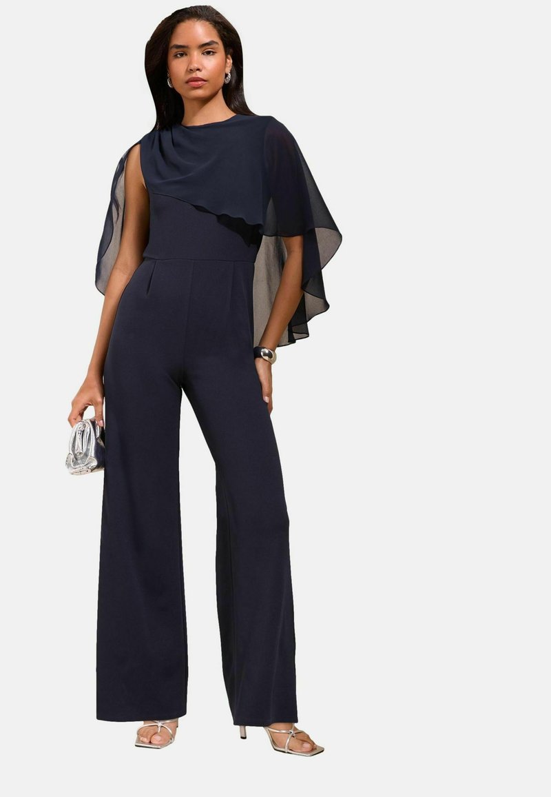 Woman wearing a navy jumpsuit with sheer cape sleeve, holding a silver clutch, wearing silver high-heeled sandals and a bracelet.