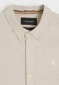 Beżowa koszula zapinana na guziki z kołnierzykiem typu button-down, kremowymi guzikami, małym emblematem lwa na piersi i czarną metką z napisem "diverse" wewnątrz kołnierza.