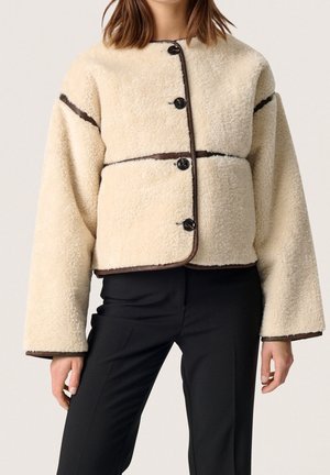 Vrouw draagt een crèmekleurig sherpa-jas met knopen en bruine accenten, gecombineerd met zwarte broek, staand tegen een effen lichte achtergrond.