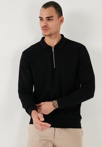 Top negro de manga larga con cuello con cremallera. Presenta una textura acanalada y puños ajustados, usado sobre pantalones beige. Diseño simple y funcional.