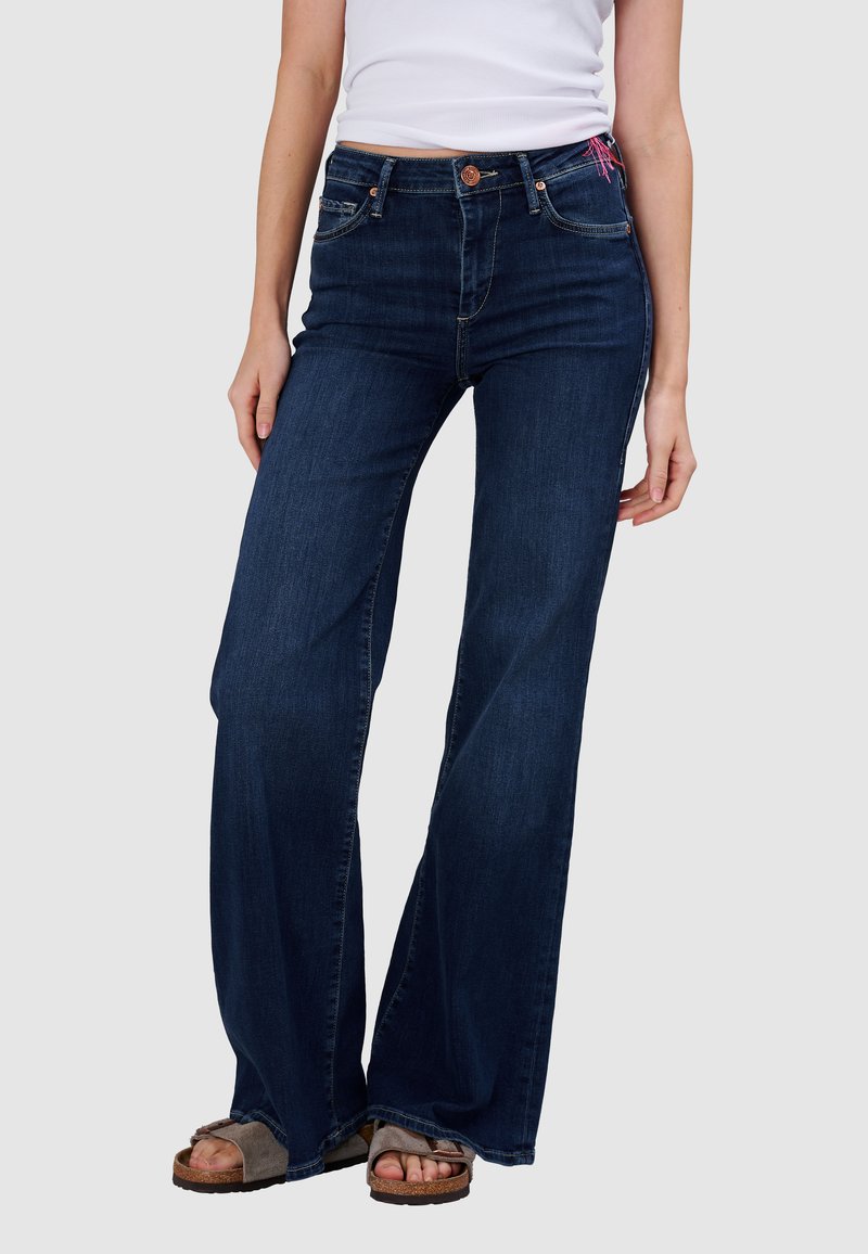 Frau trägt dunkelblaue Jeans mit weitem Bein, weißes ärmelloses Oberteil und braune Sandalen mit offenen Zehen, steht vor einem einfarbigen Hintergrund.