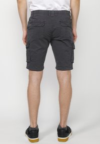 Man som bär mörkgrå cargoshorts, svarta sneakers och grå strumpor, stående v�änd bort från kameran mot en enkel ljus bakgrund.