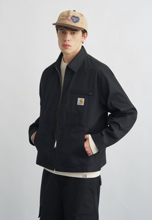 Jeune homme portant une casquette beige et une veste noire zippée, les mains dans les poches, debout devant un fond clair uni.