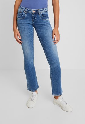 Personne portant un jean slim bleu légèrement délavé, un haut bleu rentré dedans, et des baskets blanches, debout devant un fond uni.