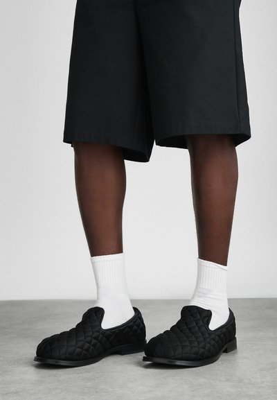 Μαύρες τσέπες slip-on παπούτσια με στρογγυλή μύτη και διακριτικές ελαστικές πλευρές, συνδυασμένα με λευκές ριγέ κάλτσες και μαύρες βερμούδες. Λείος καουτσούκ σόλα.