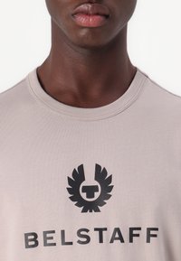 Light beige cotton t-shirt featuring a black graphic logo with wing accents in the center. Crew neck design. Smooth texture.