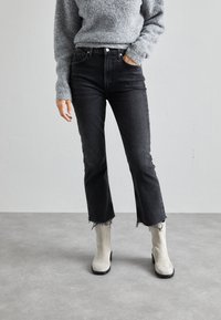 Jeans de mezclilla negros, de cintura alta, de diseño acortado y acampanado con dobladillo deshilachado, combinados con botas de ante gris claro hasta el tobillo y un suéter gris texturizado.