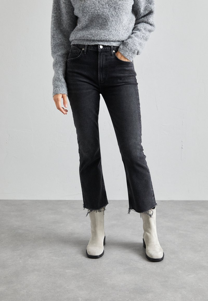 Jeans de mezclilla negros, de cintura alta, de diseño acortado y acampanado con dobladillo deshilachado, combinados con botas de ante gris claro hasta el tobillo y un suéter gris texturizado.