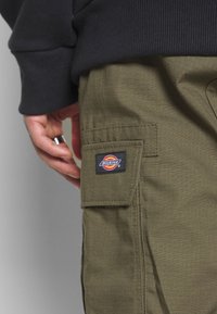 Olivgröna cargobyxor med en texturerad tyg, med en sidficka och en svart Dickies-logotyp patch. Handen vilar på midjebandet.