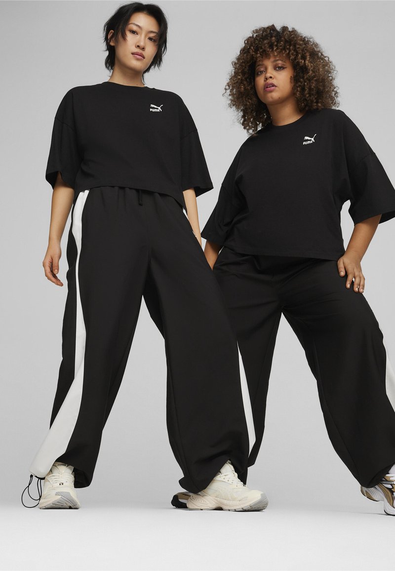 Zwarte oversized T-shirts en zwarte wijde broek met witte zijstrepen, gedragen door twee modellen, gecombineerd met lichtgekleurde sneakers.