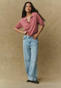 Rožnato obarvan kratek top z rokavi v kombinaciji s svetlo modrimi kavbojkami širokih hlačnic. Kavbojke imajo rahlo oprane robove. Model stoji z roko na boku.