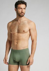 Zielone bokserki wykonane z gładkiego materiału, o dopasowanym kroju i elastycznej talii, prezentowane na męskim modelu na neutralnym tle.