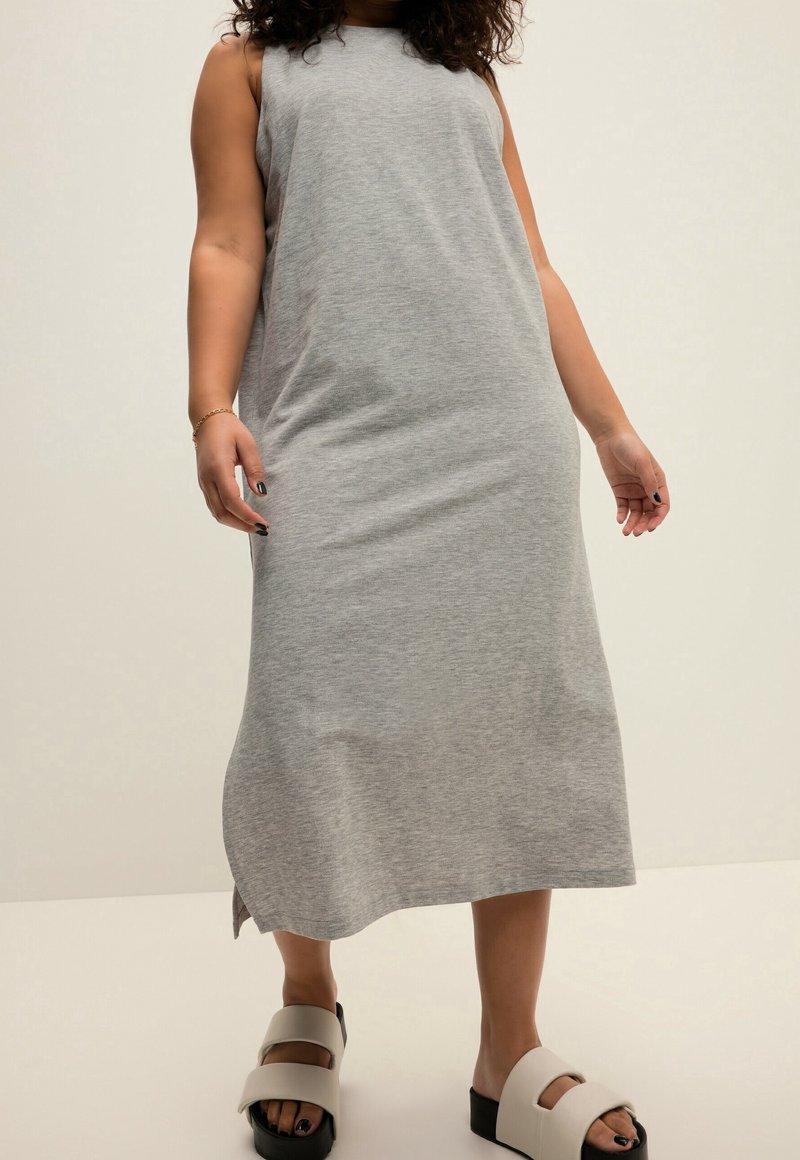 Grijze, mouwloze maxi-jurk met een losse pasvorm, zachte textuur en zijsplitten. Gecombineerd met brede zwarte en witte slip-on sandalen.