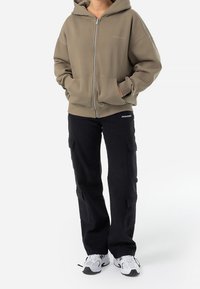Sudadera con capucha y cremallera en verde oliva, combinada con pantalones cargo negros que tienen bolsillos laterales. El calzado incluye zapatillas blancas con acentos grises.