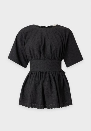 Femme portant un top péplum noir à manches courtes, un short beige, des sandales noires, des boucles d'oreilles créoles dorées et un collier, debout devant un fond clair uni.