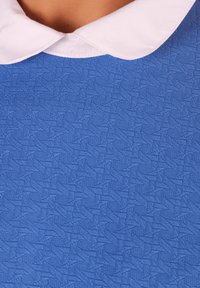 Tejido azul texturizado con un patrón geométrico, con un cuello blanco. La textura parece ligeramente elevada, añadiendo interés visual.