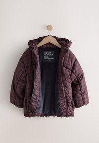 Doudoune matelassée à carreaux rouges et bleu marine avec capuche, dotée d'une fermeture éclair et d'une doublure intérieure douce et sombre. Suspendue à un crochet en bois.