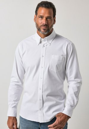 Mand med skæg iført en hvid skjorte med knapper og brystlomme samt blå jeans, stående mod en ensfarvet lys baggrund.
