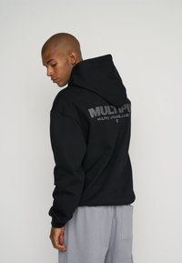 Black hooded sweatshirt with large grey text on the back, featuring a relaxed fit and ribbed cuffs, paired with light grey pants.