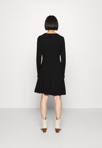 Vestido longo preto com mangas, com uma saia rodada, feito de um tecido suave. Apresenta um decote redondo e uma textura lisa. Usado com botas de tornozelo de cor clara.
