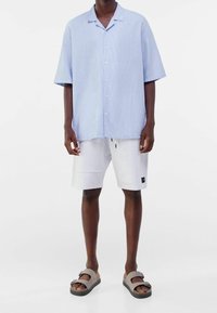 Světle modrá košile s krátkým rukávem a vertikálním proužkovaným vzorem, se zapínáním vpředu; spárovaná s bílými šortkami a šedými skládacími sandály.