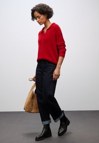 Roter gestrickter Pullover mit Kragen, dunkelblaue gerade Jeans, schwarze Knöchelstiefel und eine beigefarbene Wildleder-Tasche. Strukturierter Stoff und umgeschlagene Ärmel sind sichtbar.
