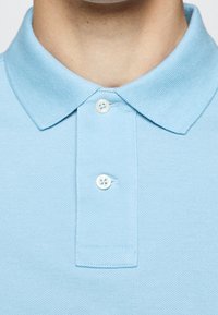 Ljusblå pikékrage med två vita knappar knäppta vid en persons hals och övre bröstområde.