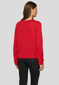 Rot gestrickter Pullover mit langen Ärmeln, rundem Halsausschnitt und lockerer Passform. Kombiniert mit dunklen Jeans, die eine strukturierte Oberfläche zeigen.