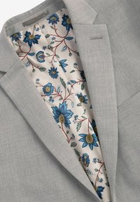 Helle graue Blazer mit glatter Textur. Das Innenfutter zeigt ein florales Muster in Blau und Beige auf einem cremigen Hintergrund. Auffälliges Reversdetail sichtbar.