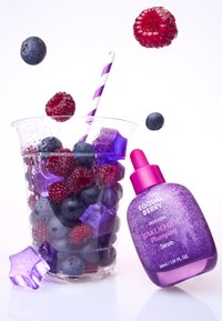 Przezroczysty plastikowy kubek z jagodami, malinami i fioletowymi kostkami lodu. Fioletowa butelka serum z pipetą, na białym tle, z paskowanym słomką.