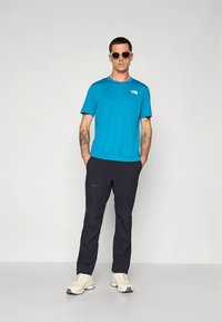 Camiseta de manga curta azul brilhante, calças pretas com bolsos, material leve, usada com óculos de sol e sapatos claros.