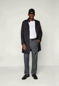 Črni plašč čez belo srajco s pisanjem, sivi hlače in črne čevlje. Dodatki vključujejo temne sončne očala in črno kapo.