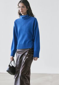 Woman wearing a blue turtleneck sweater and brown leather skirt, holding a small black handbag, walking against a plain background.